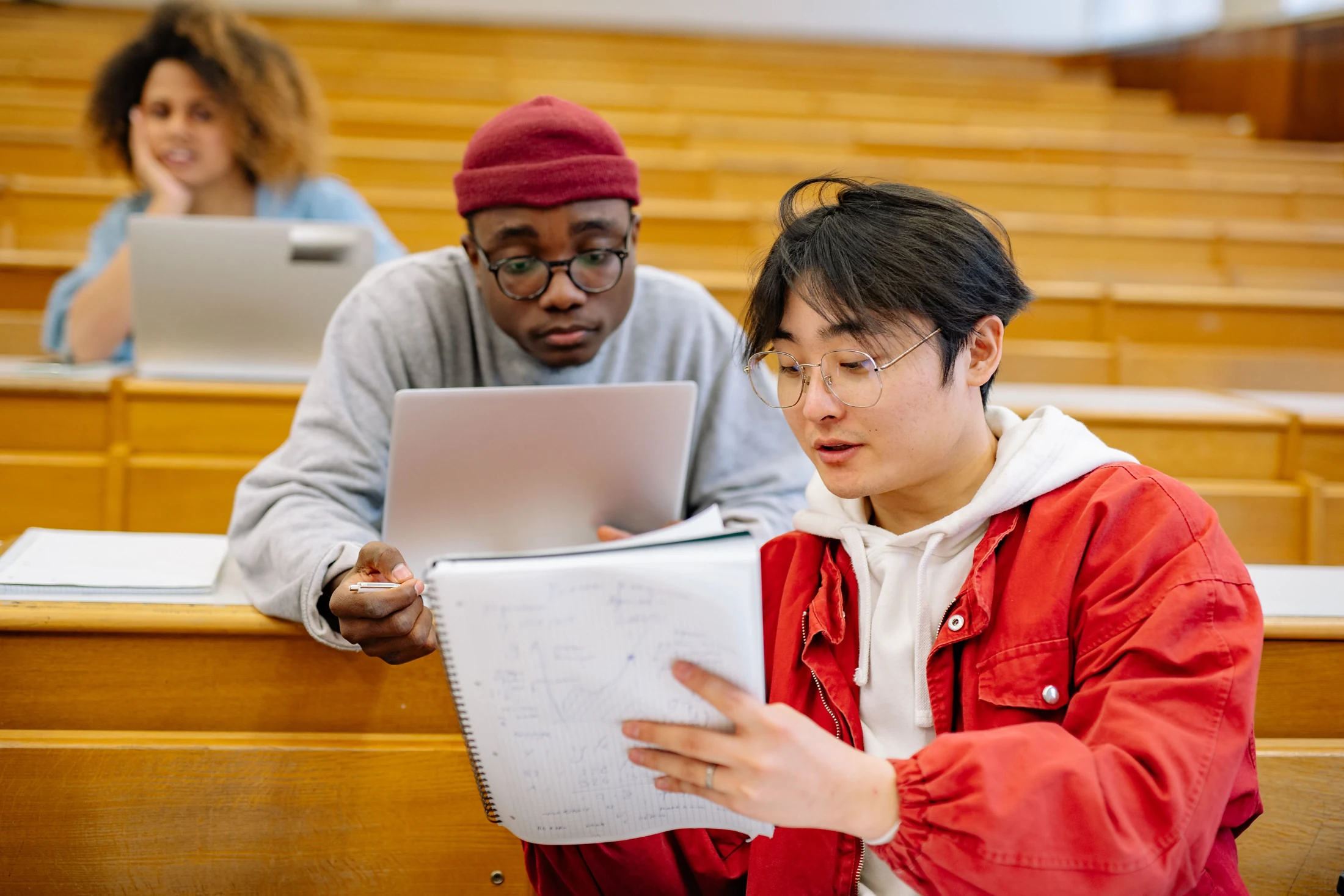 A student shows notes to a classmate with a laptop in a university lecture hall.
