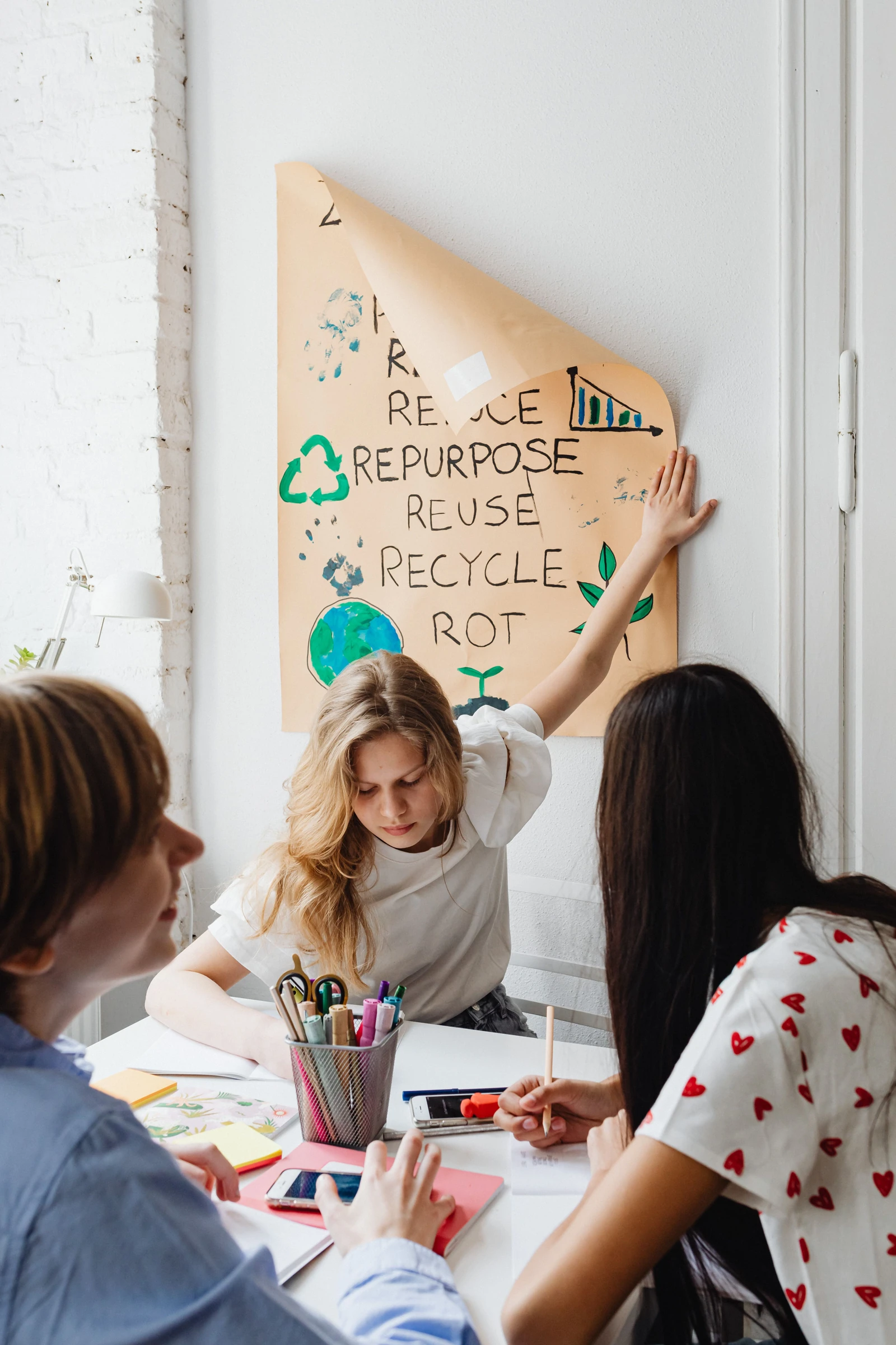 Three students work on a school project with paper, markers and a recycling poster.